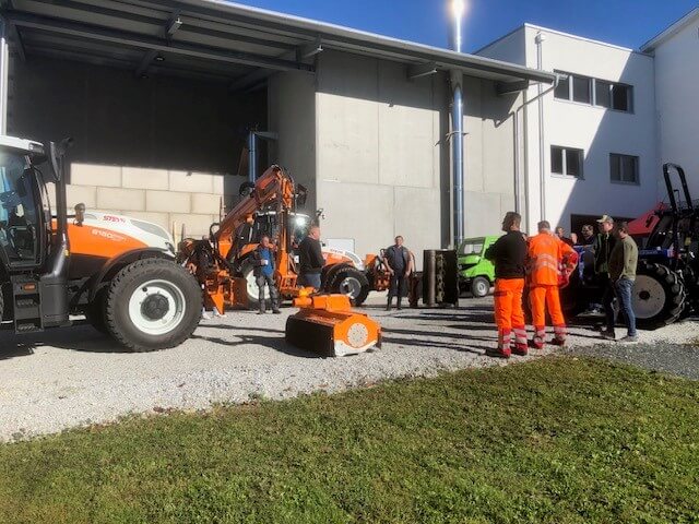 Mitarbeiter besichtigen orange Baumaschinen im Freien bei sonnigem Wetter vor einem Industriegebäude.