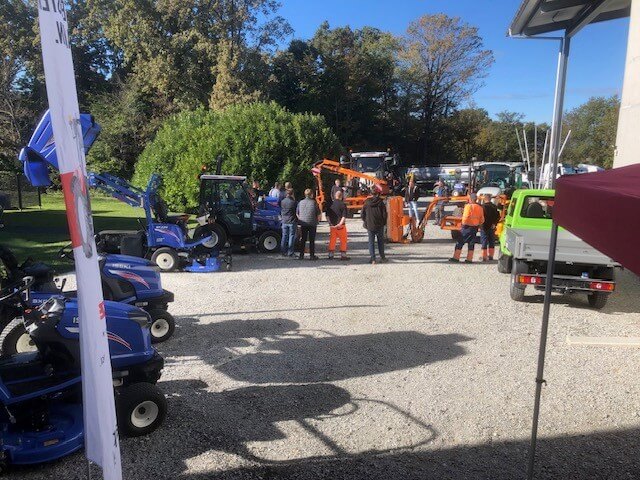 Outdoor-Ausstellung von Landwirtschaftsmaschinen mit Besuchern bei sonnigem Wetter und grüner Kulisse.