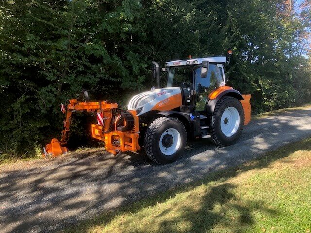 Traktor mit Anbaugerät auf Waldweg bei Sonnenschein, umgeben von grünen Bäumen. Ideal für Landschaftspflege und Forstarbeit.