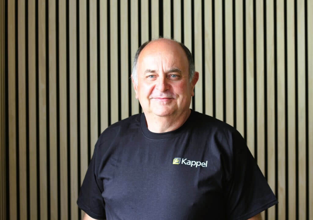 Ein lächelnder Mann in einem Kappel T-Shirt vor einer Holzlamellenwand.