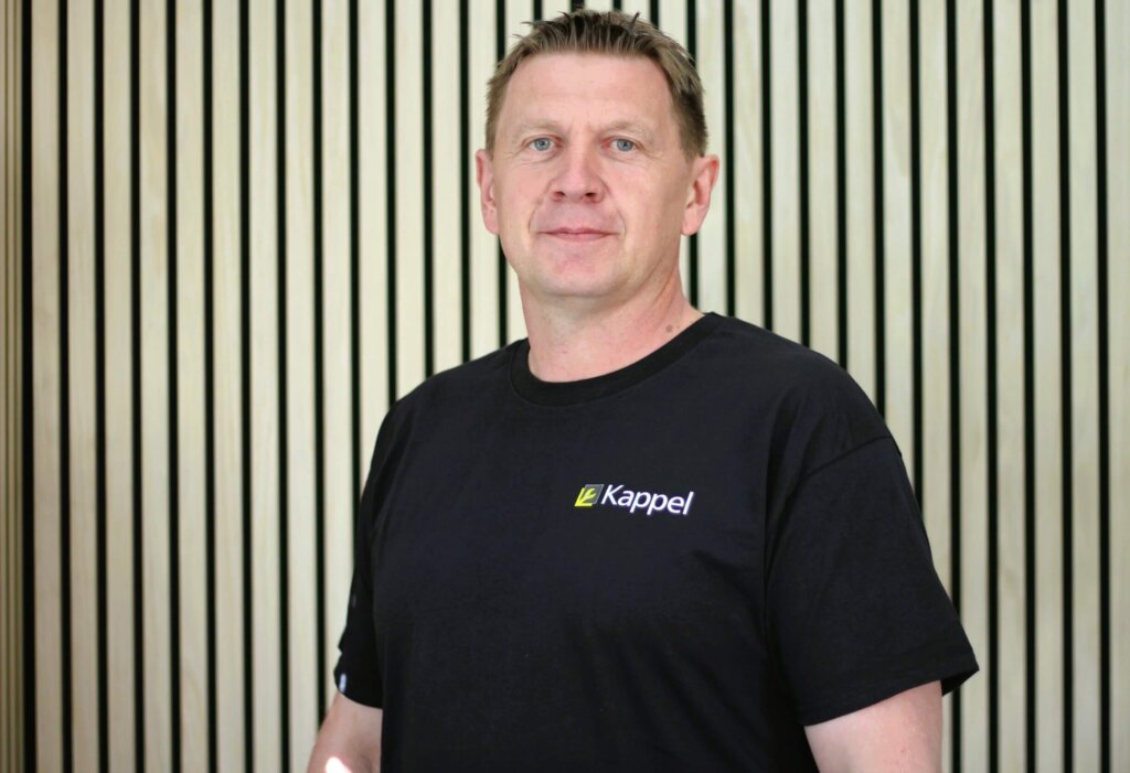 Mann im schwarzen T-Shirt steht vor einer Wand mit Holzverkleidung. Kappel-Logo auf dem T-Shirt.