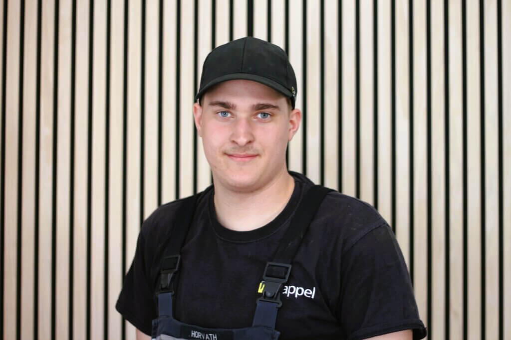 Junger Mann mit Baseballmütze und schwarzem T-Shirt vor gestreiftem Hintergrund, schaute freundlich in die Kamera.