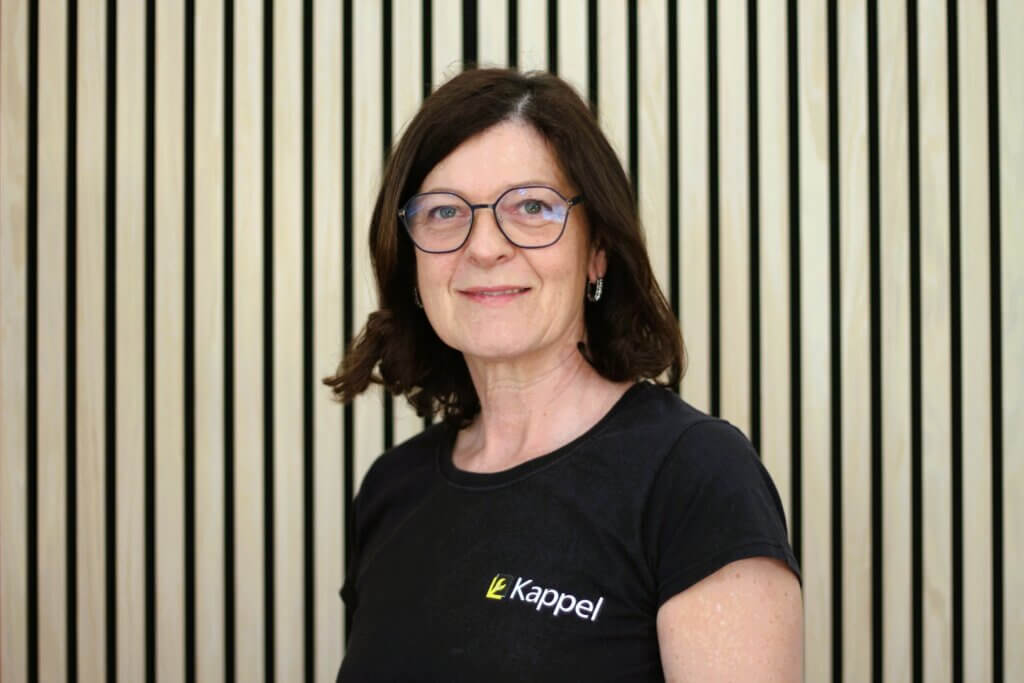 Eine Frau mit Brille und schwarzem T-Shirt steht vor einer Holzlattenwand und lächelt in die Kamera.