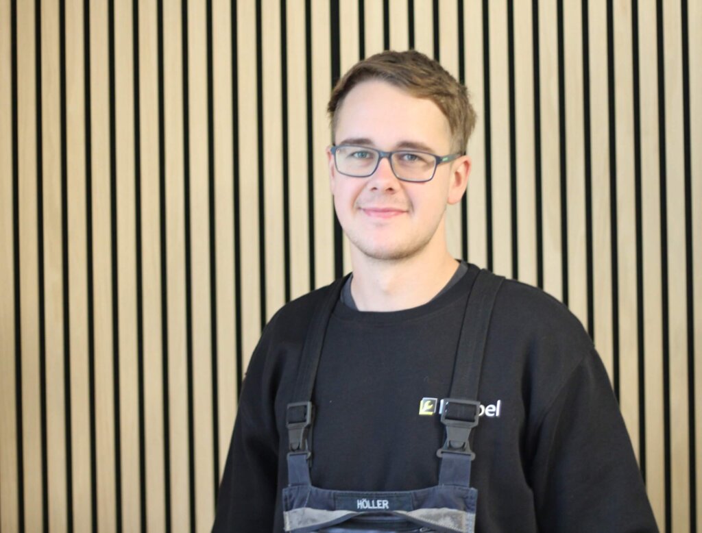 Junger Mann in Arbeitskleidung vor Streifenwand, freundlich lächelnd und Brille tragend.