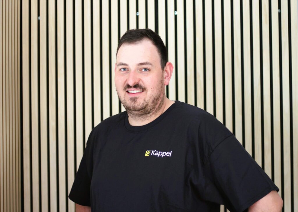 Mann im schwarzen T-Shirt mit Kappel-Logo vor Holzlamellenwand, freundlich lächelnd.
