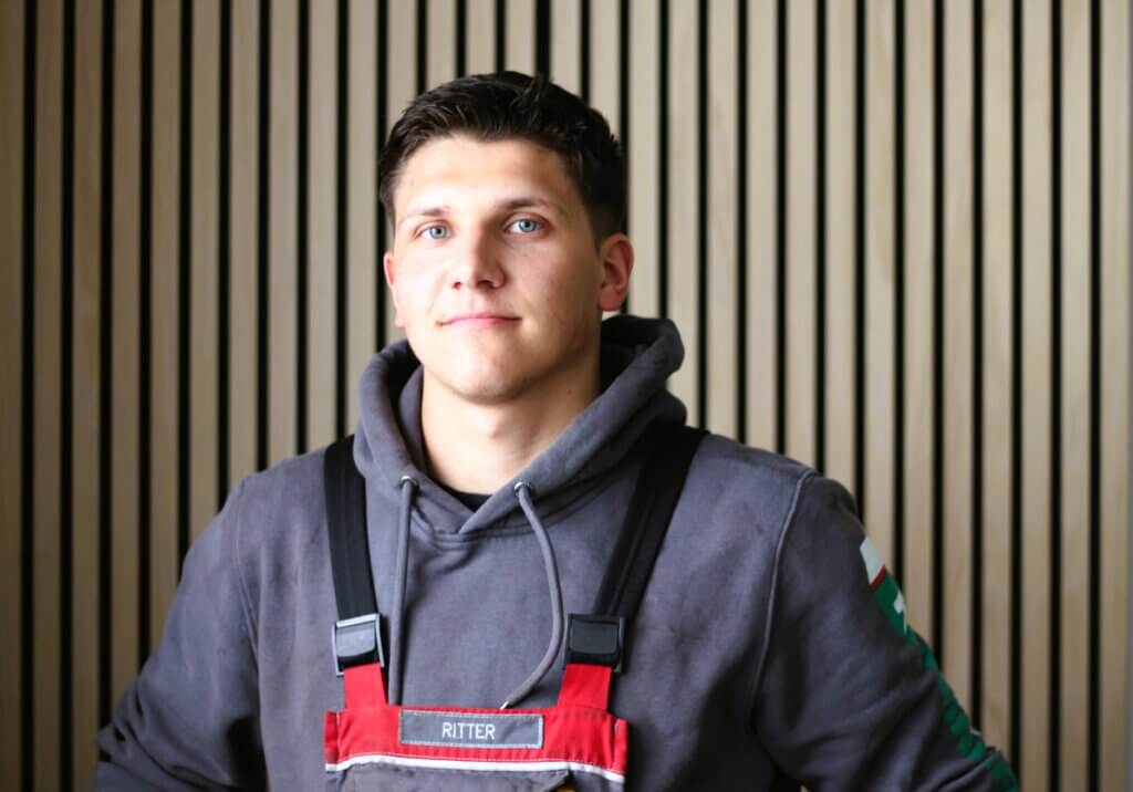Junger Mann in grauem Hoodie und roter Arbeitsschürze vor einer gestreiften Holzwand.