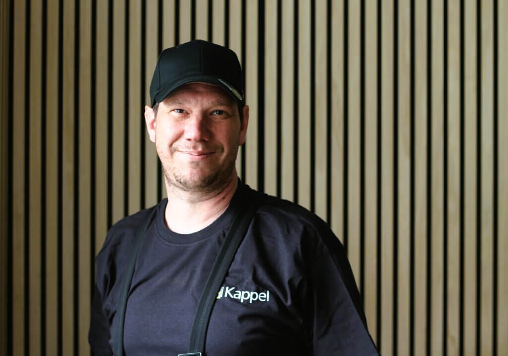 Ein lächelnder Mann in schwarzem T-Shirt und Cap vor einem Hintergrund aus Holzlatten.