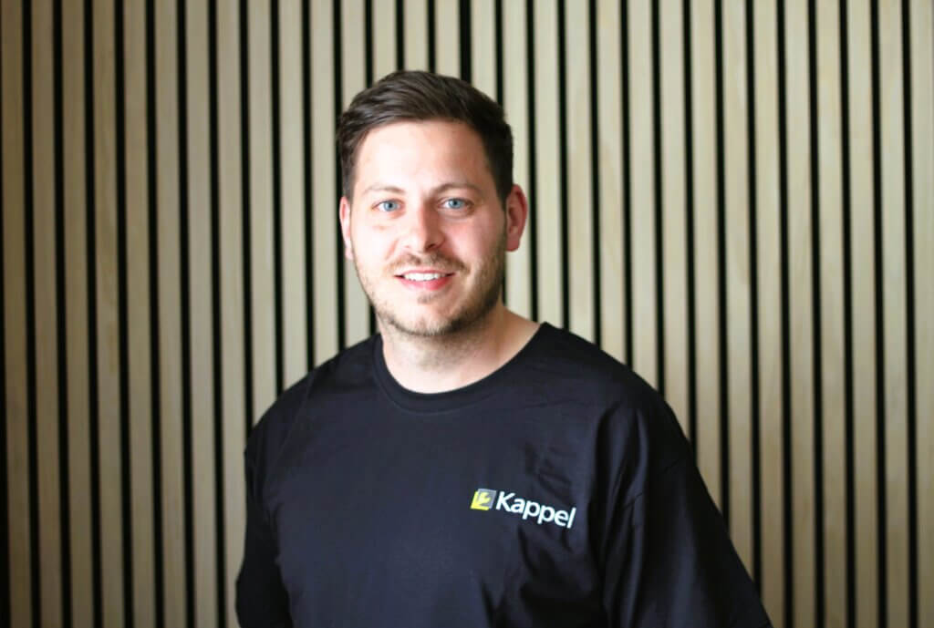 Ein lächelnder Mann in einem schwarzen Kappel-T-Shirt vor einem Holzpaneel-Hintergrund.