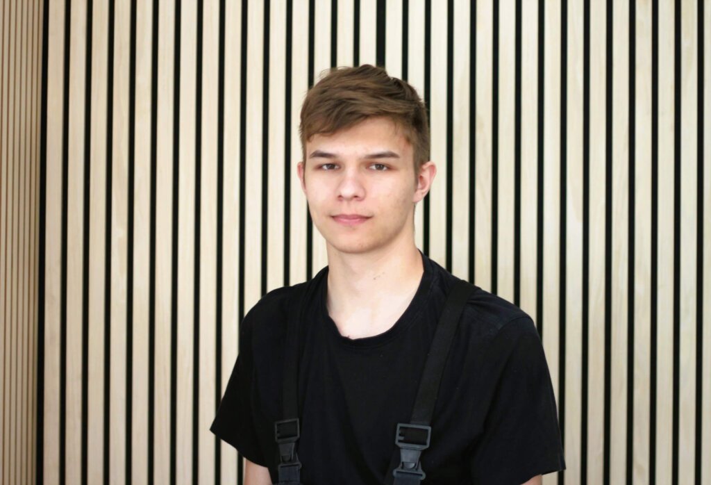 Junger Mann in schwarzem T-Shirt vor elegantem, gestreiftem Holzhintergrund.