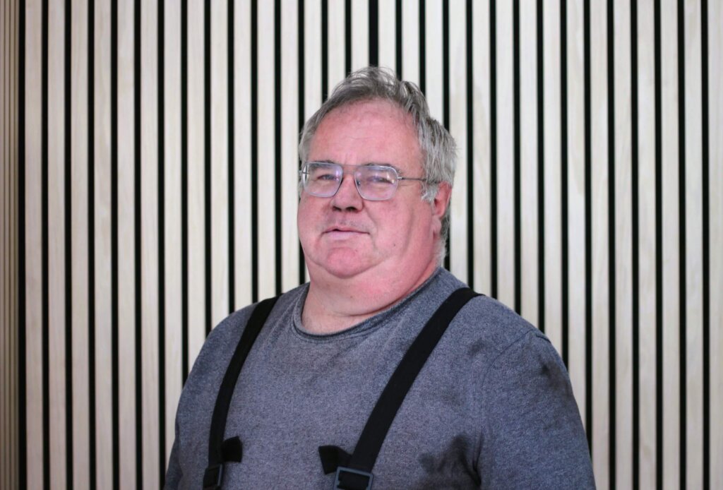 Älterer Mann mit Brille und grauem Pullover vor einem gestreiften Holzwand-Hintergrund.