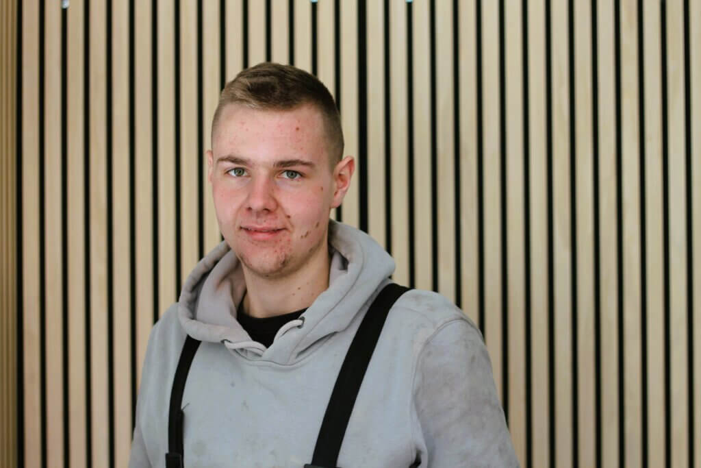 Junger Mann in grauem Hoodie vor Holzwand, lächelt in die Kamera. Perfekt für Handwerker- oder DIY-Themen.