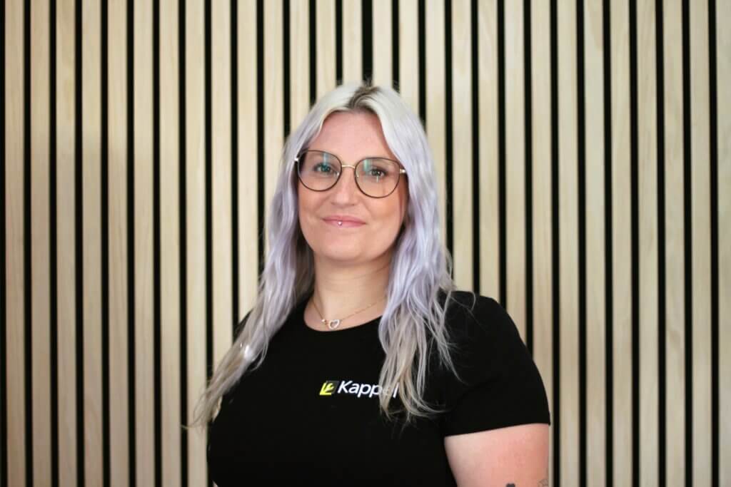 Frau mit Brille und langen, blonden Haaren vor modernen Holzpaneelen, trägt ein schwarzes Oberteil.