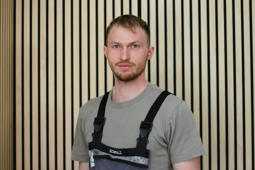 Junger Mann in Arbeitskleidung vor moderner Holzwand, blickt freundlich in die Kamera.