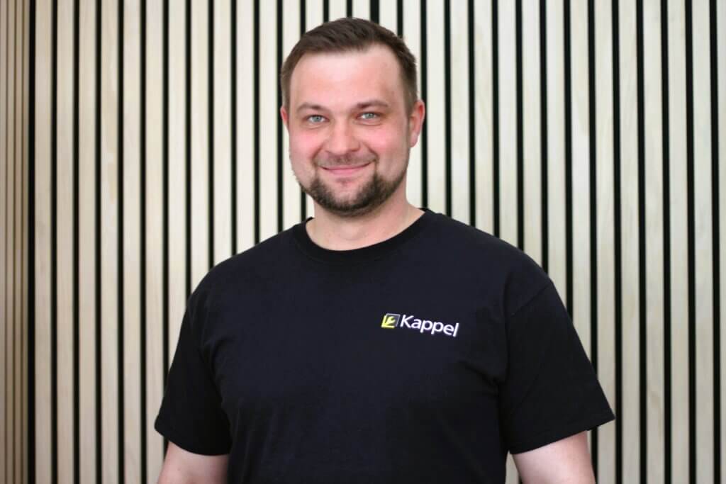 Lächelnder Mann im Kappel-T-Shirt vor Holzlattenwand. Optimaler Alt-Text für Firmenwebsites oder Teamseiten.