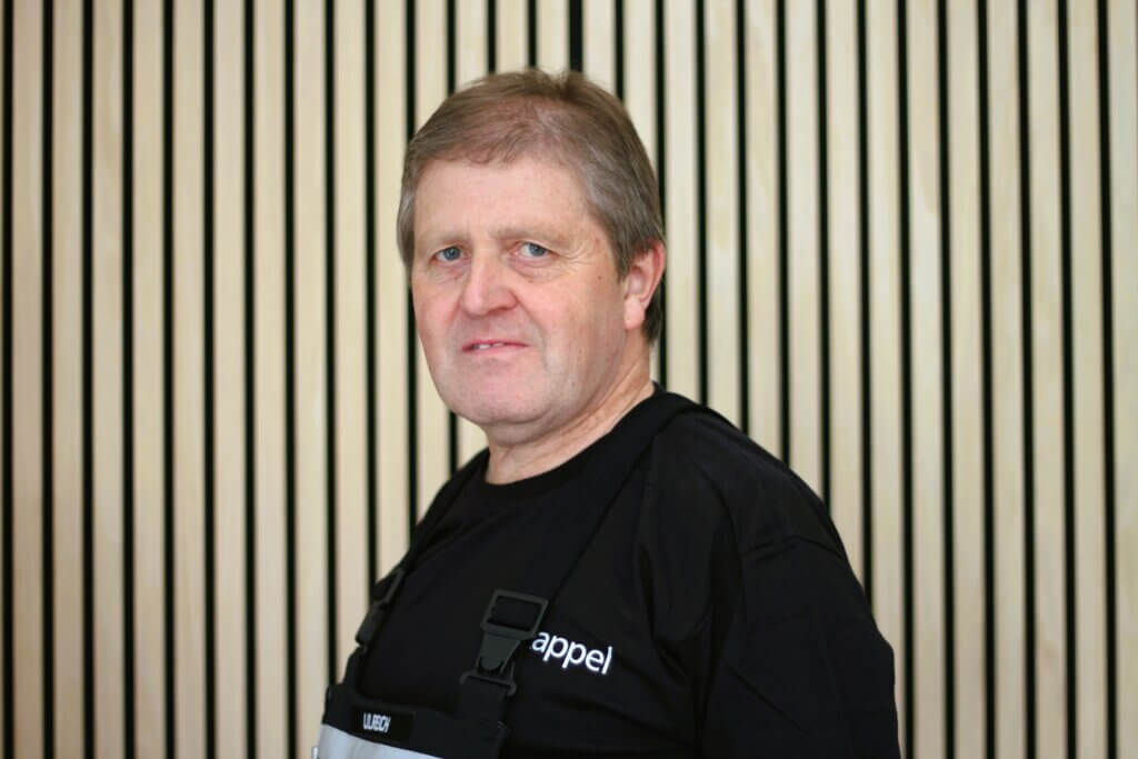 Mann in schwarzem Shirt vor gestreifter Holzwand, lächelt leicht in die Kamera.