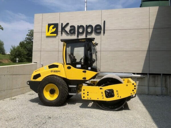 Gelber Straßenwalzenverdichter von BOMAG vor einem Gebäude mit Kappel-Logo, seitliche Ansicht auf Kiesfläche.