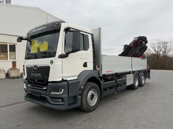 Weißer MAN-Lkw auf einem Parkplatz mit Kranaufbau von Schwarzmüller im Hintergrund einer grauen Industrieumgebung.