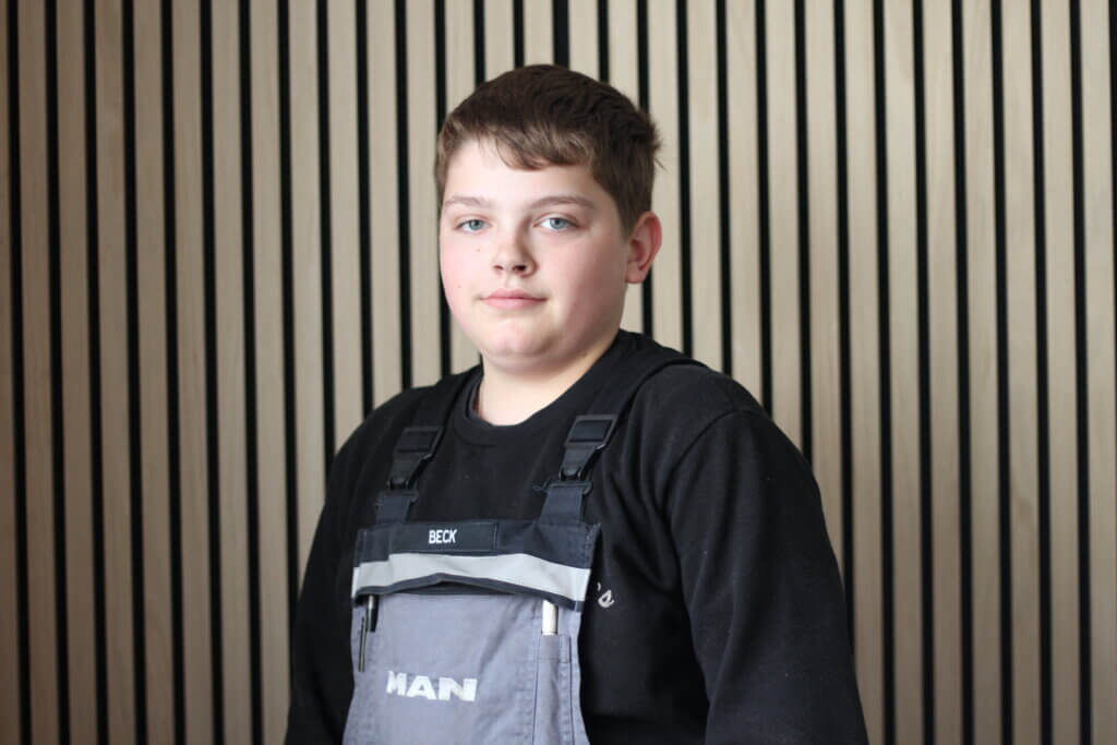 Junger Mechaniker in Arbeitskleidung vor Holzlamellenwand.