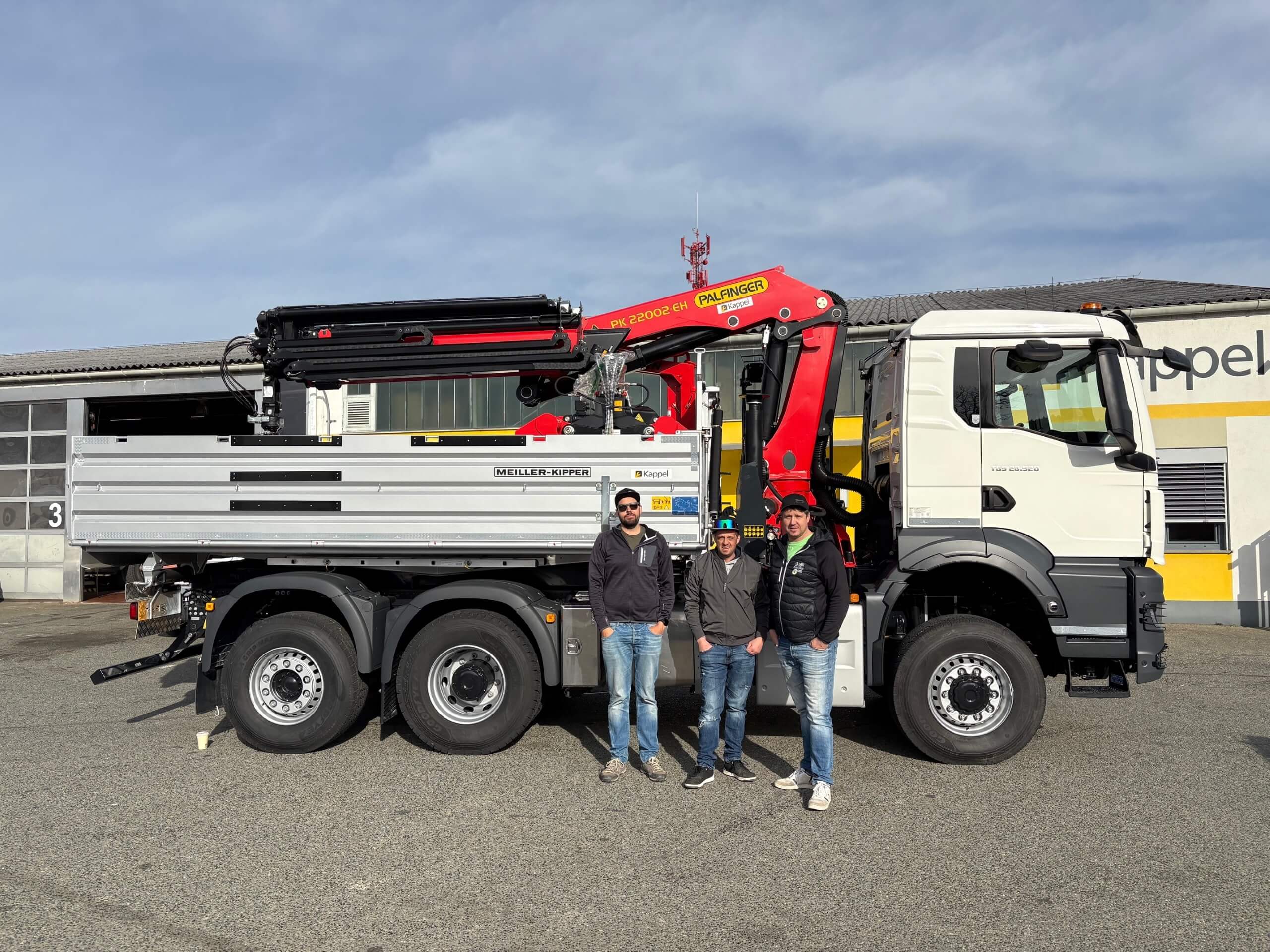 LKW mit Palfinger-Kran vor Werkstatt, drei Personen stehen davor, blauer Himmel im Hintergrund.