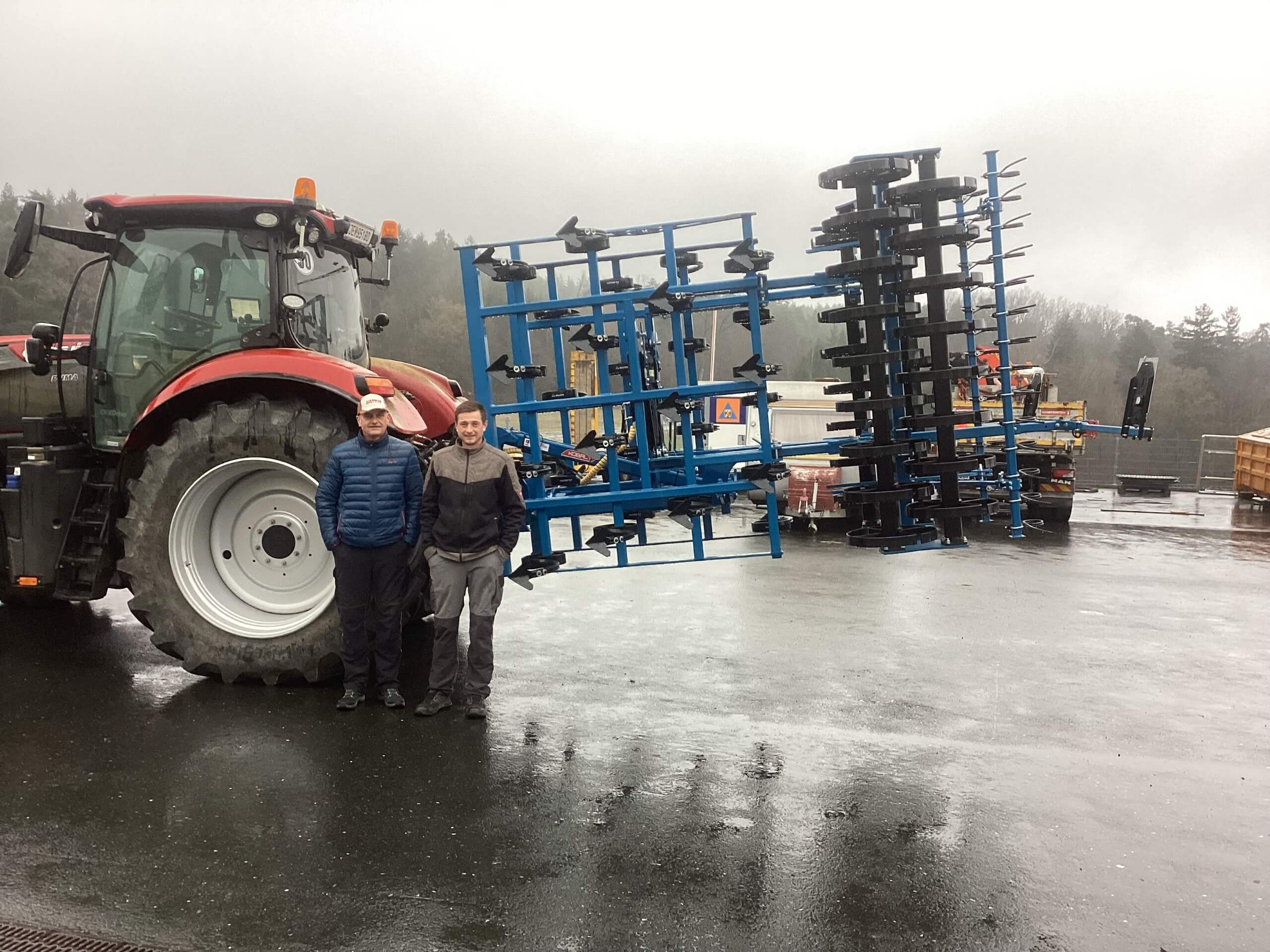 Traktor mit landwirtschaftlichem Gerät und zwei Personen auf einem Nieselregenplatz vor bewölktem Himmel.