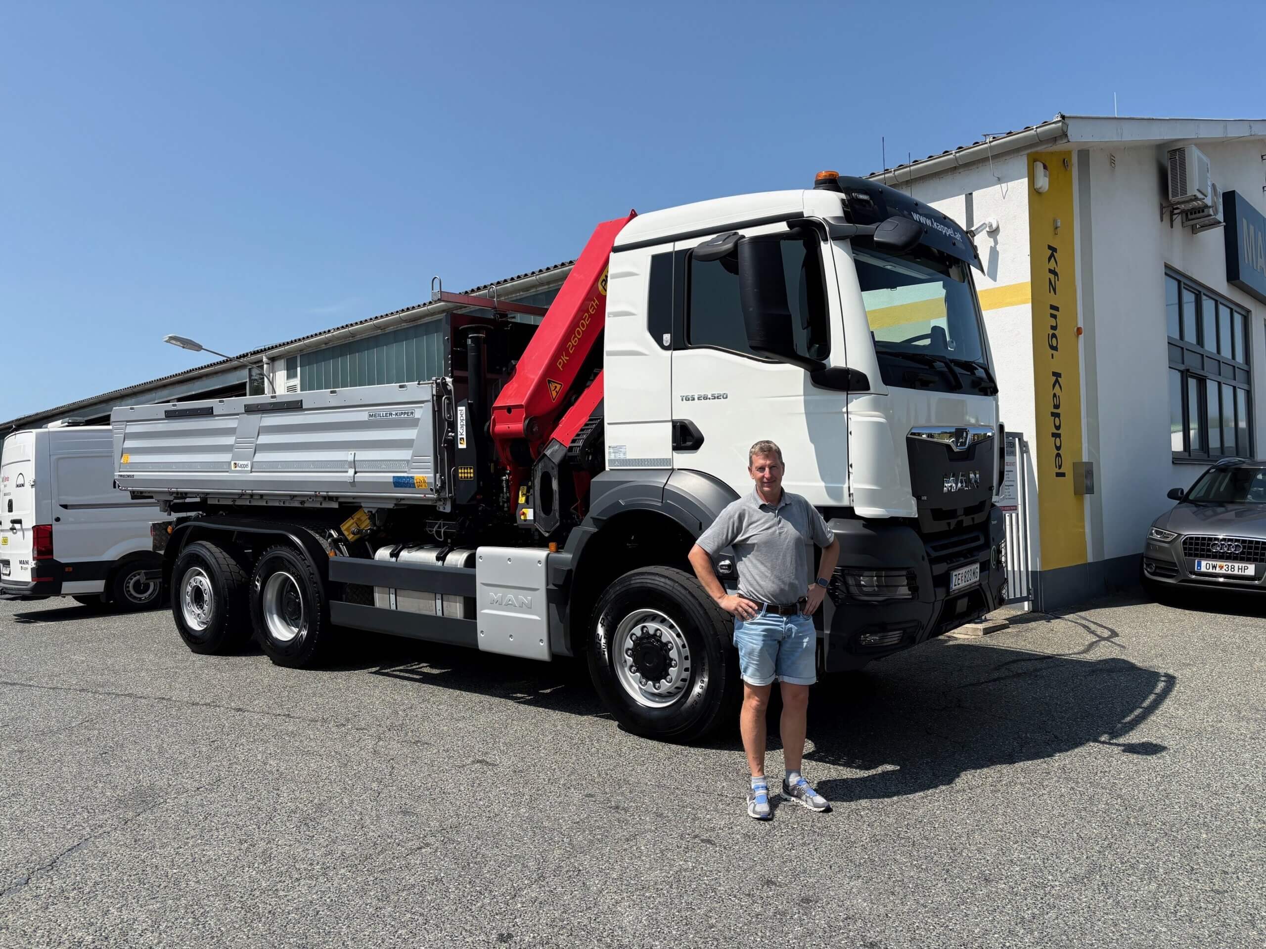 Mann steht vor MAN-LKW auf Parkplatz vor Werkstattgebäude bei sonnigem Wetter.