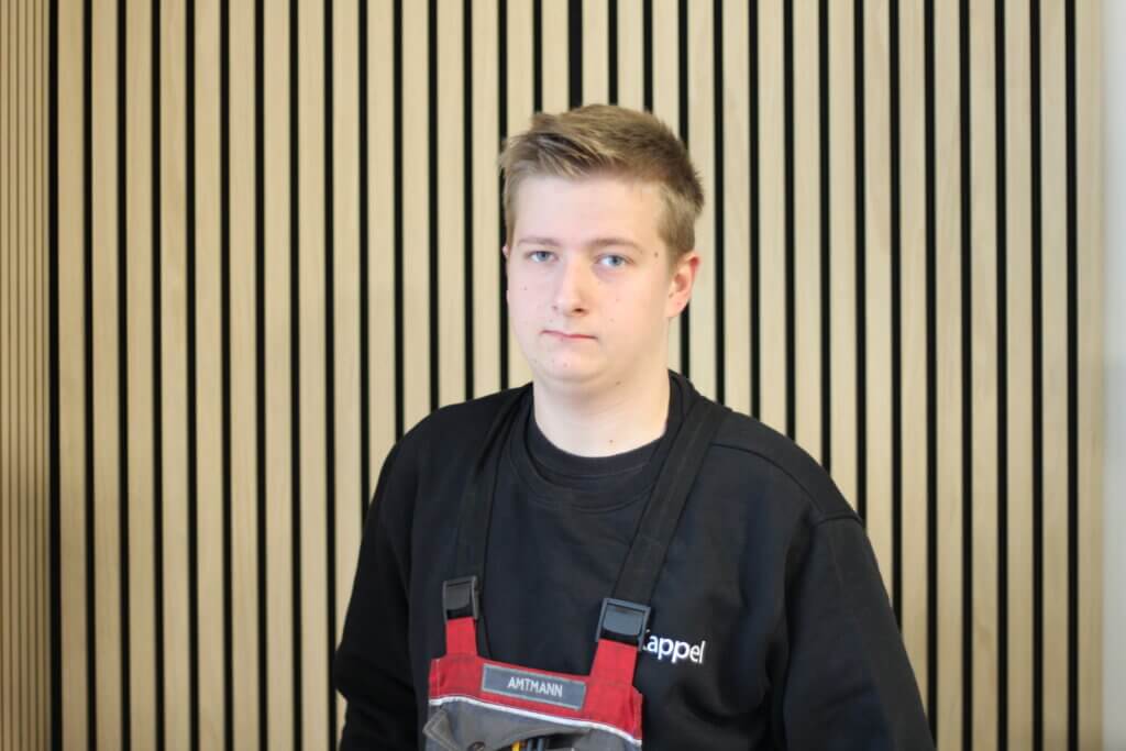 Junger Handwerker in Arbeitskleidung vor Holzlattenwand.