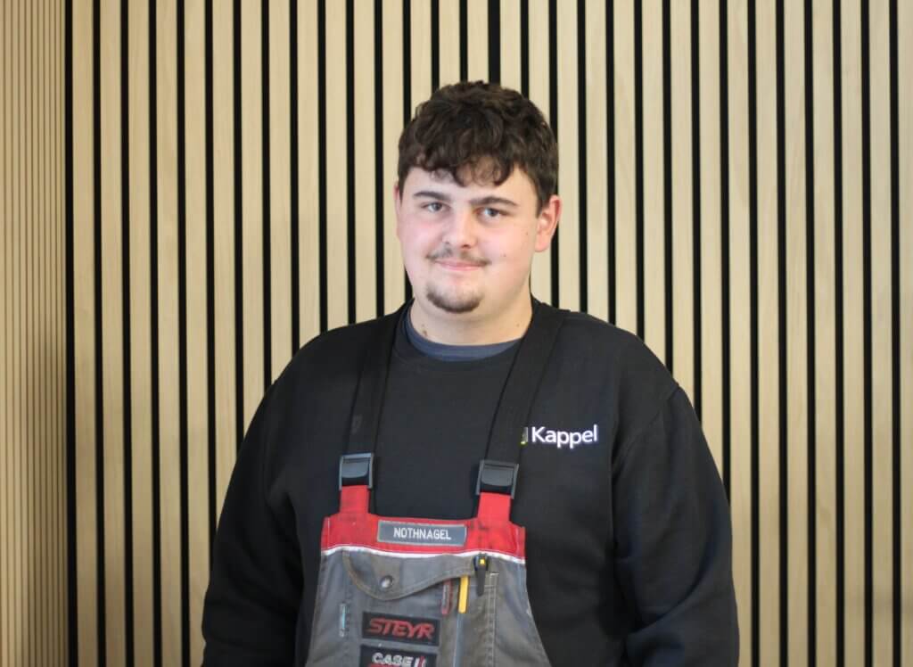 Junger Mann in Arbeitskleidung steht vor einer Wand mit vertikalen Holzpanelen und schaut in die Kamera.