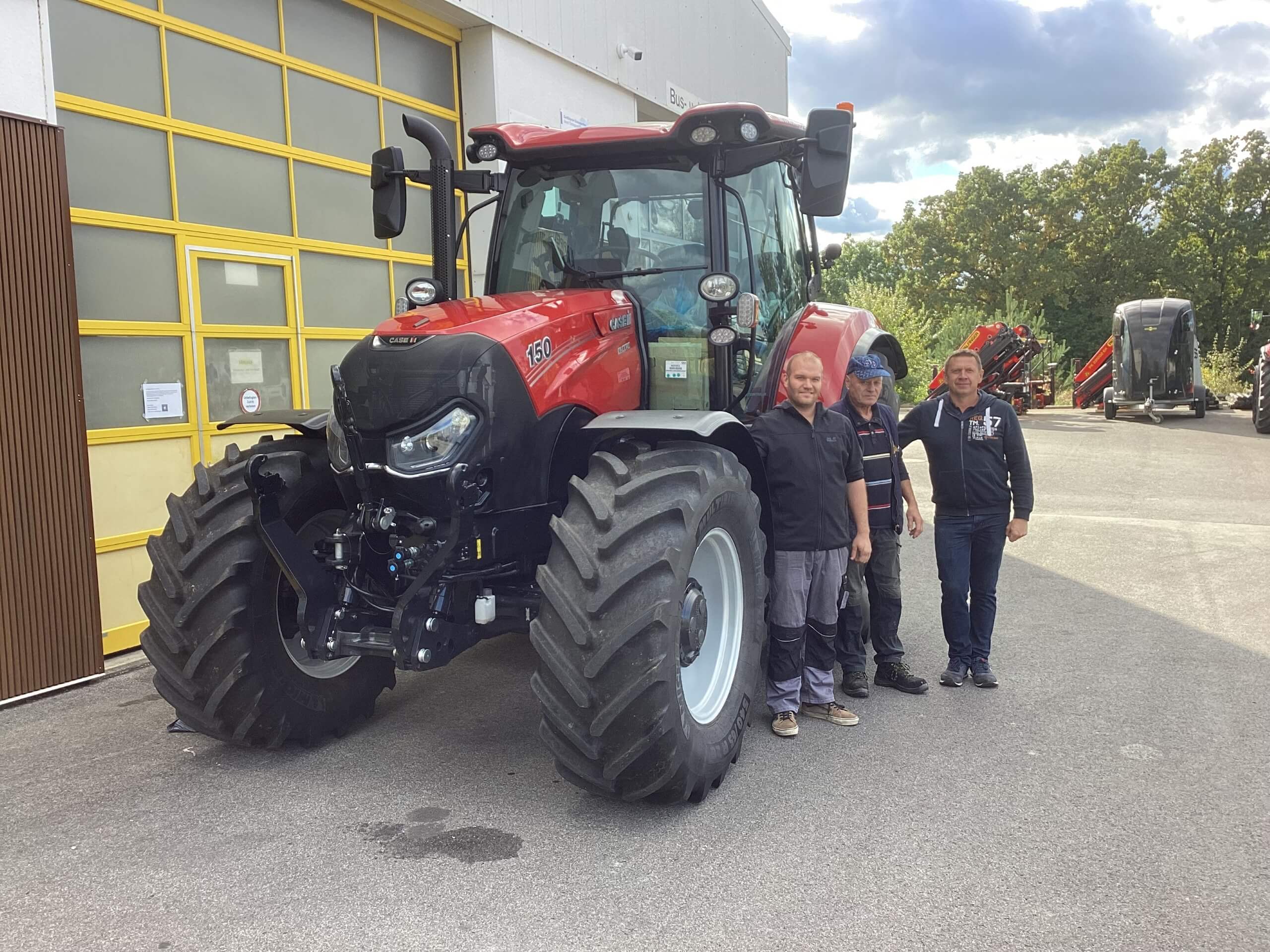 Drei Männer stehen neben einem großen, roten Traktor vor einer Werkstatt mit gelbem Tor.
