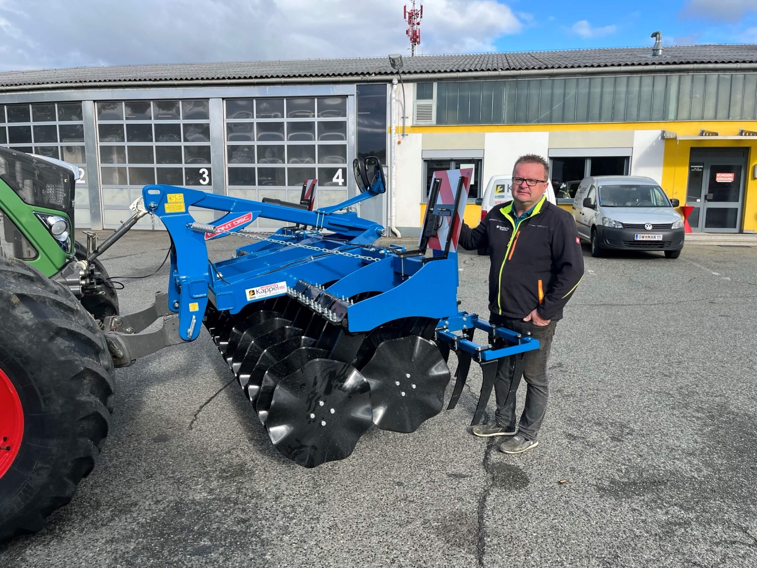 Mann steht neben einem blauen Landmaschinen-Anbaugerät vor einer Lagerhalle.