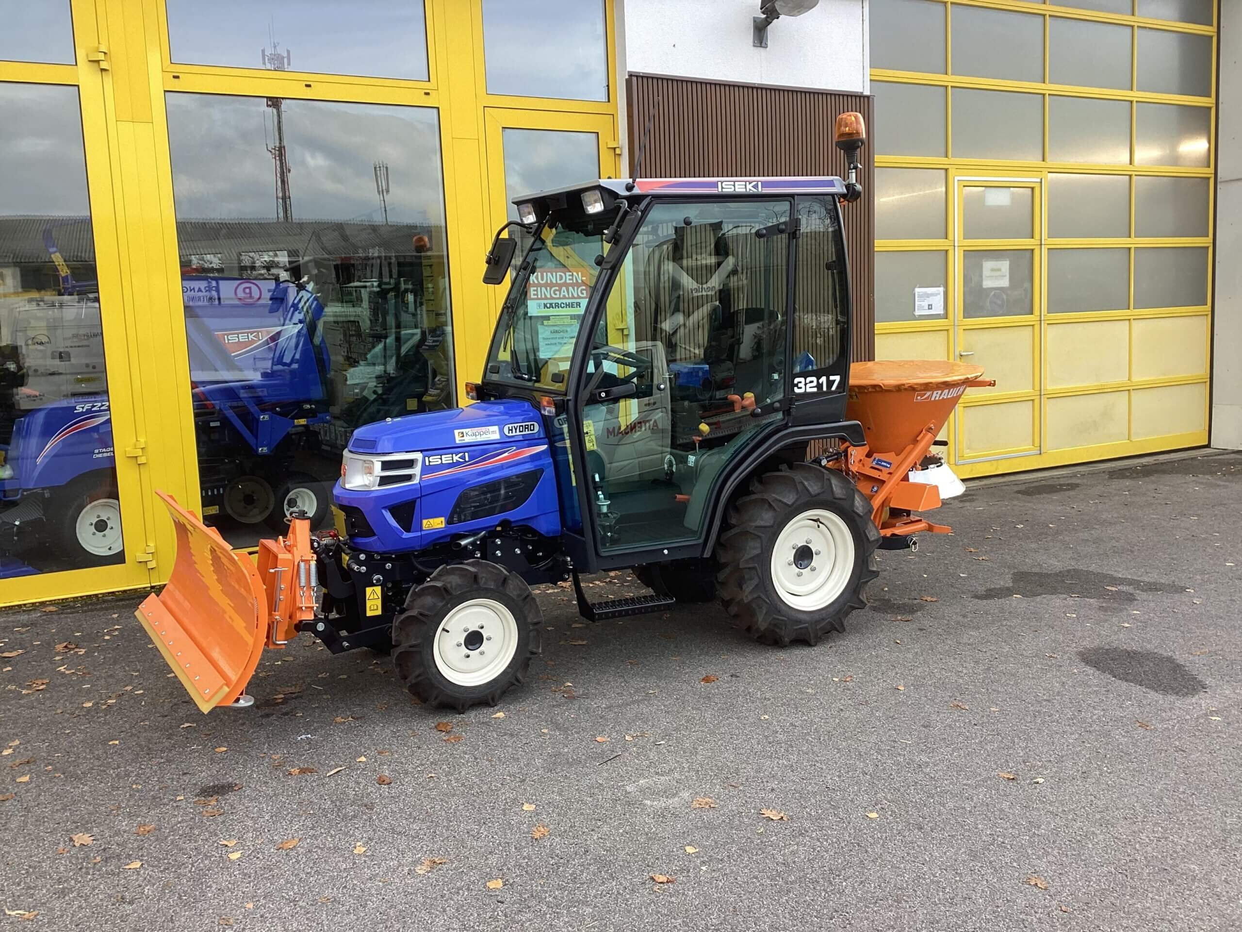 Kompakter blauer Traktor mit Schneepflug und Streuer, vor gelber Werkstatt geparkt. Ideal für Winterdienst.