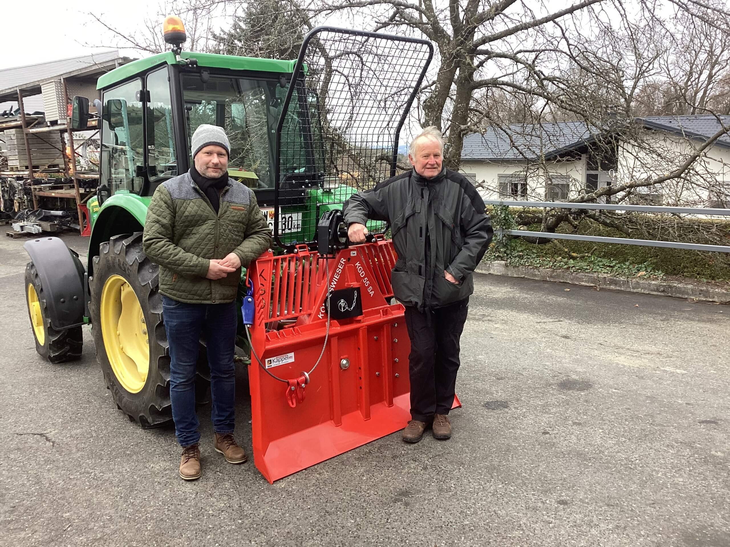 Zwei Männer posieren mit einem Traktor und einem roten Anbaugerät auf einem Bauernhof im Freien.