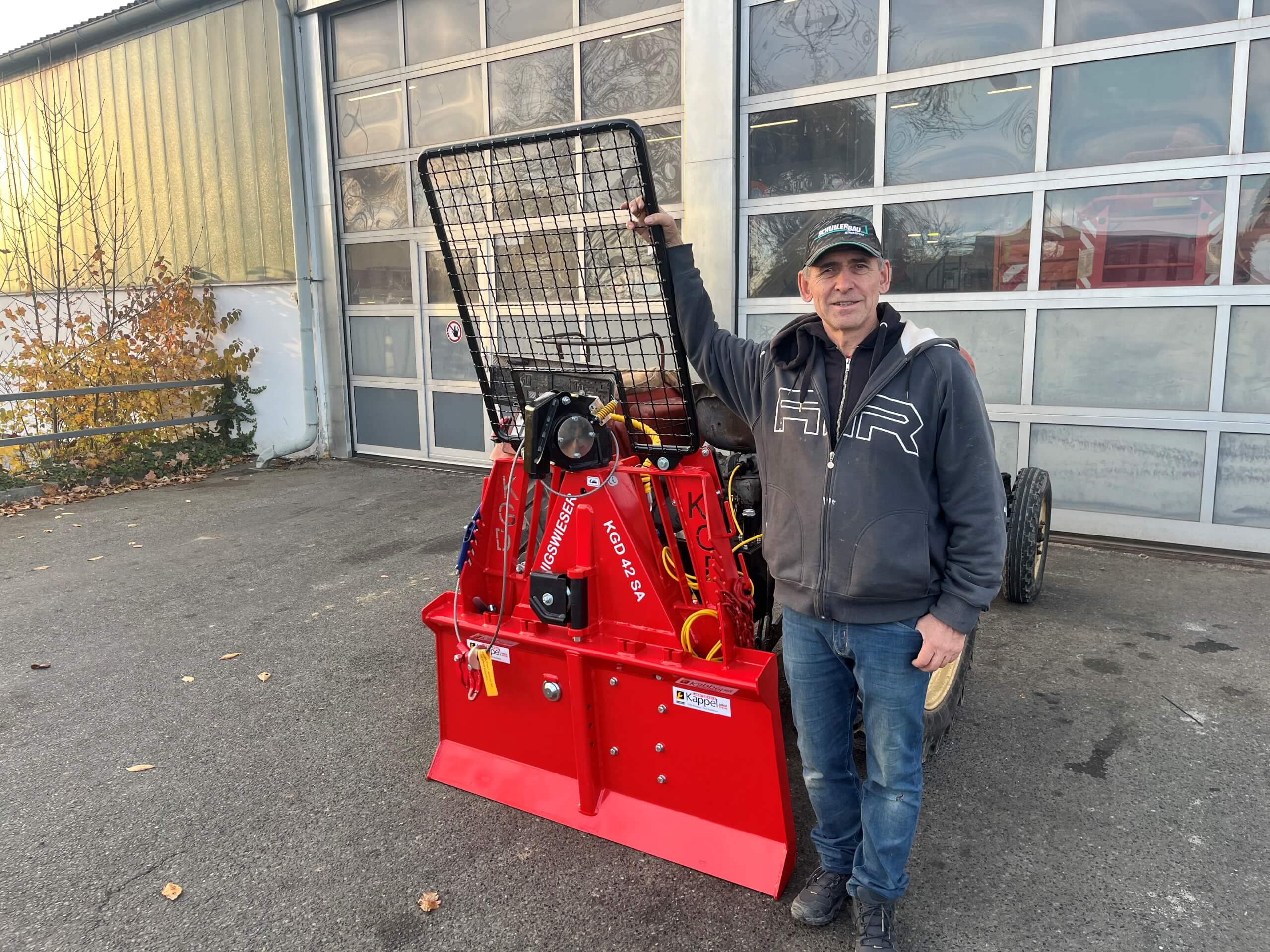 Mann neben roter Forstseilwinde vor Garagentor im Freien, Herbstlaub im Hintergrund.