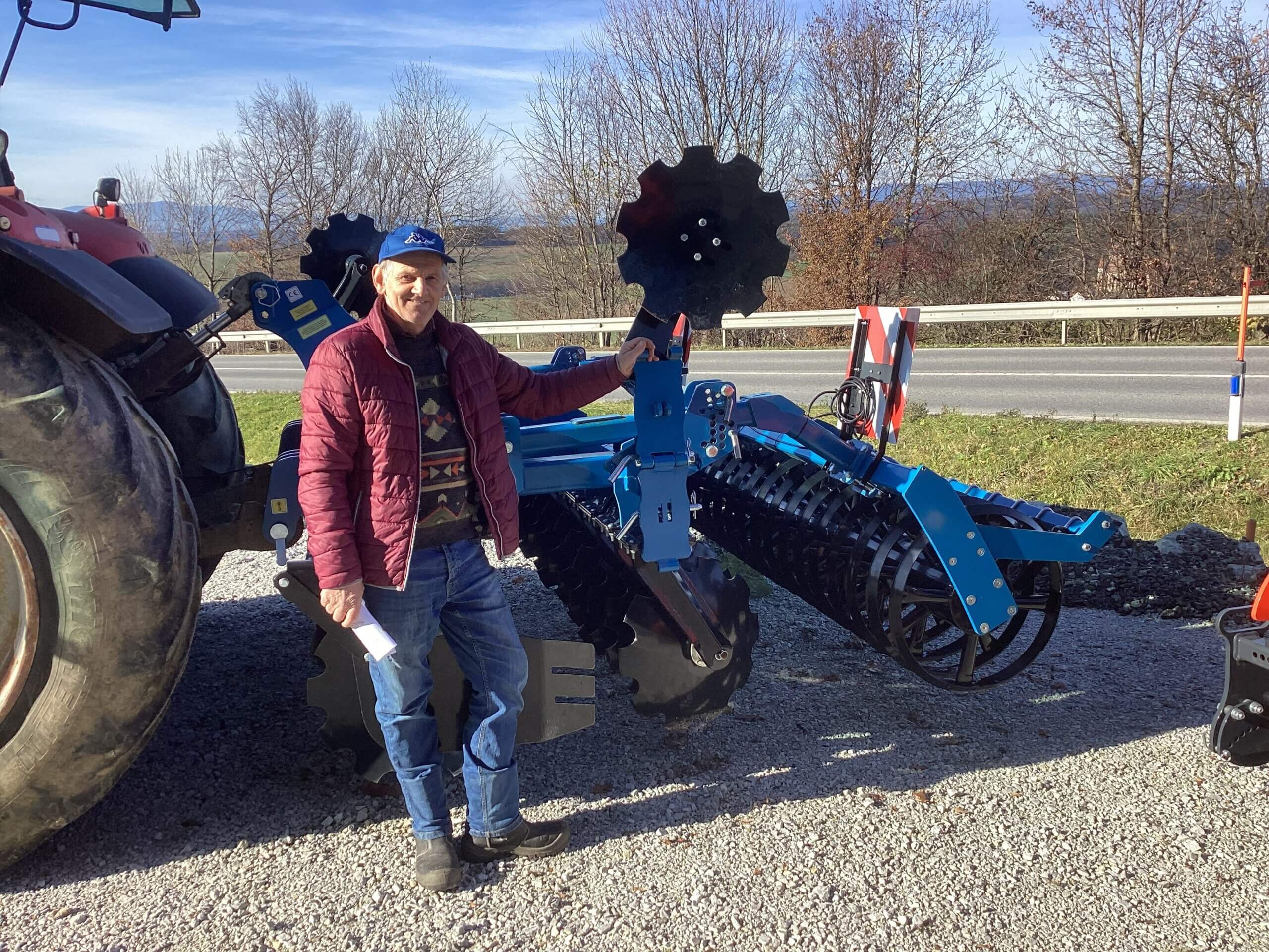 Mann steht neben einem blauen landwirtschaftlichen Gerät vor einem Traktor im Freien an einem sonnigen Tag.