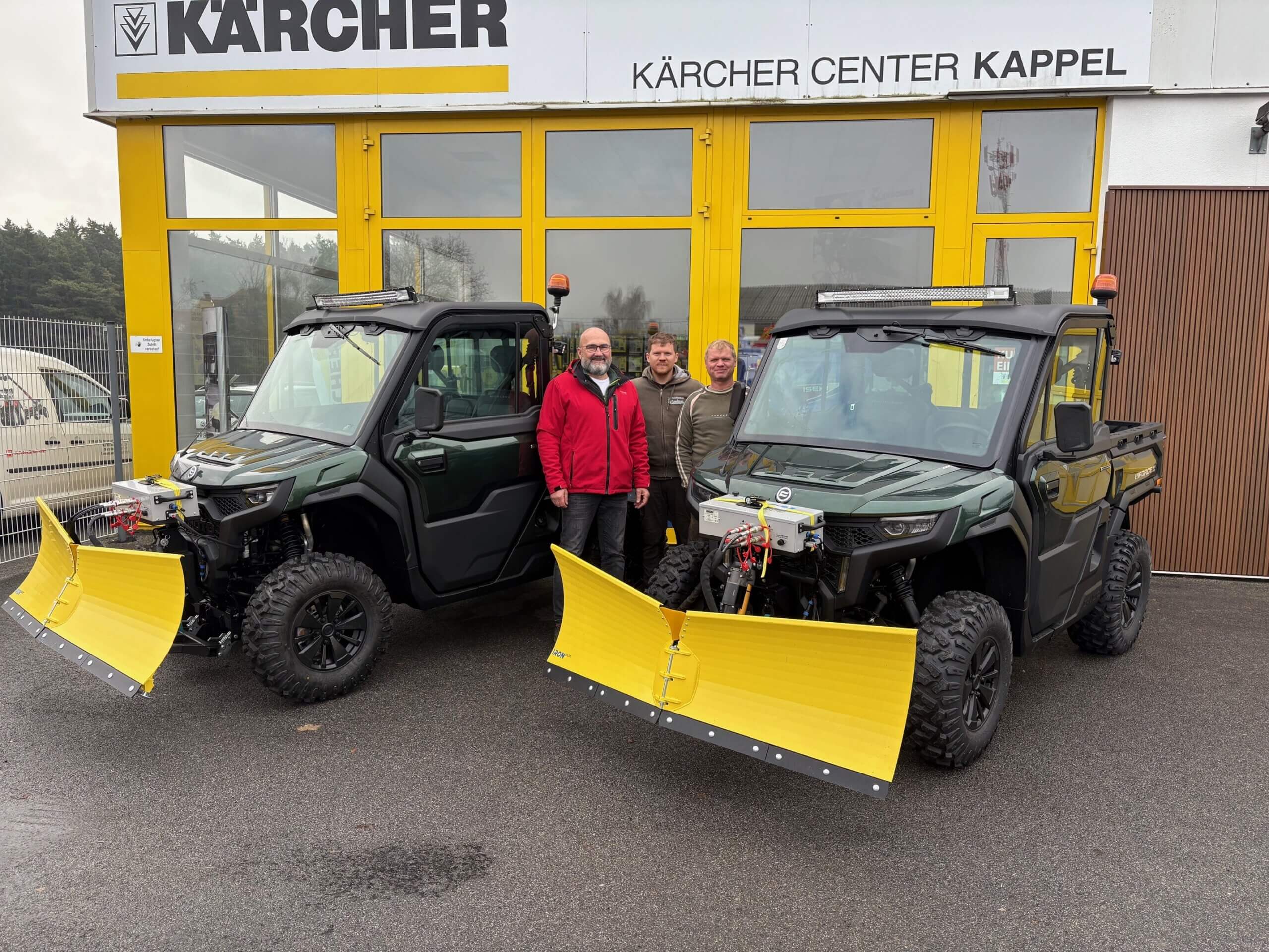 Kärcher Center Kappel: Zwei grüne Schneepflüge mit gelben Schaufeln und vier Personen vor dem Gebäude.