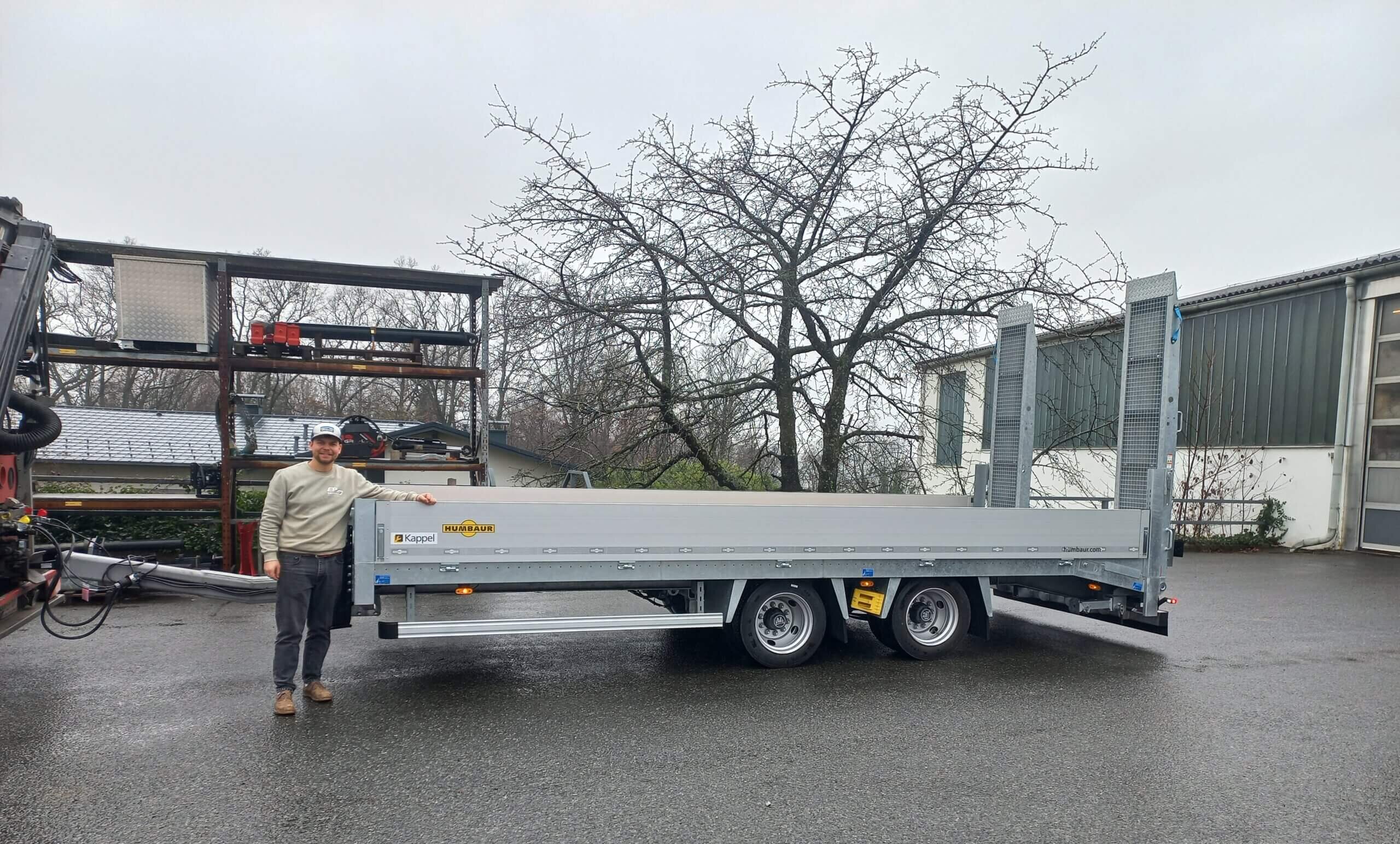 Mann posiert neben Humbaur Anhänger auf Parkplatz, grauer Himmel, Bäume im Hintergrund. Ideal für Transport und Logistik.