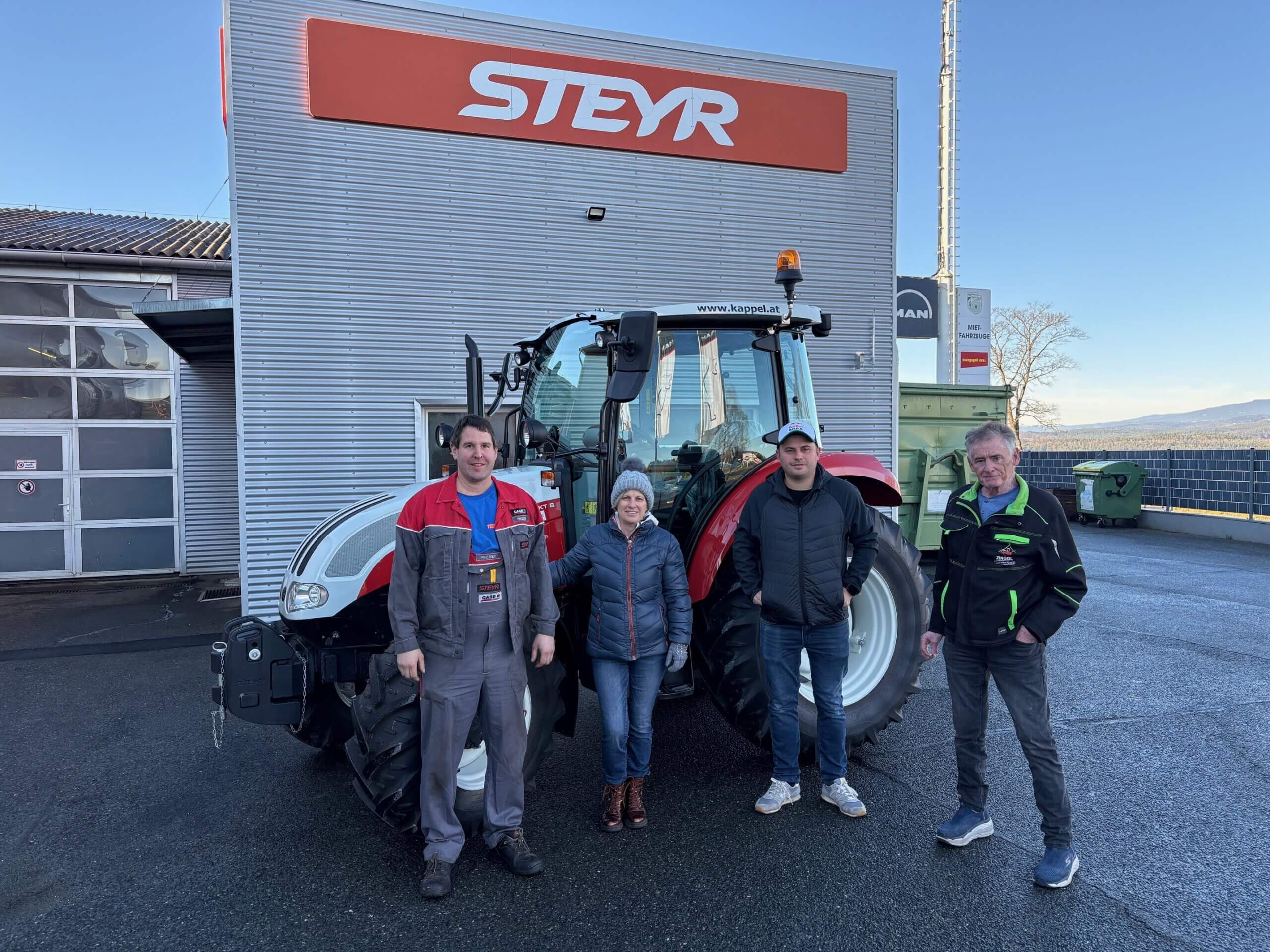 Vier Personen vor einem Steyr-Traktor außerhalb eines Werkstattgebäudes an einem sonnigen Tag.