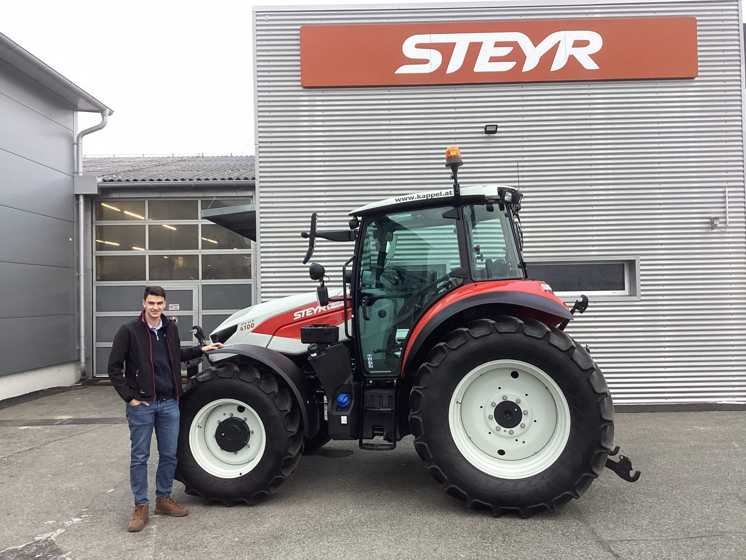 Mann steht neben einem Steyr Traktor vor einem Gebäude mit Steyr-Logo. Optimal für landwirtschaftliche Arbeiten.