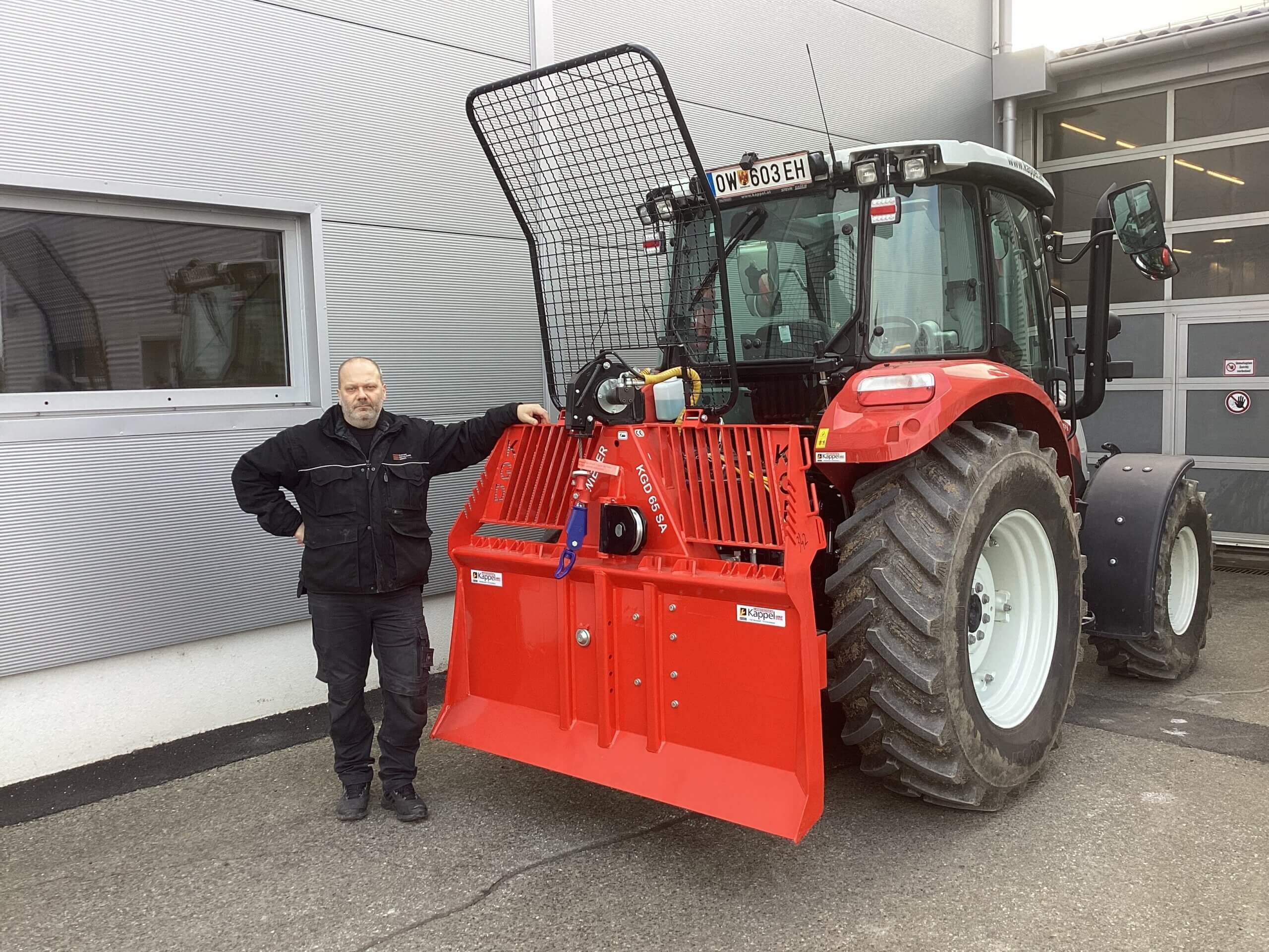 Person neben rotem Traktor mit forstwirtschaftlicher Ausrüstung, vor Industriegebäude geparkt.