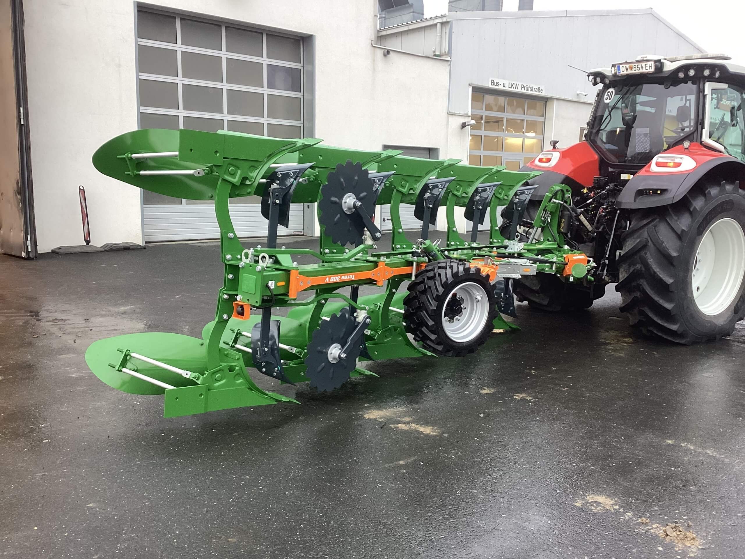 Moderner Traktor mit grünem Pflug auf nassem Hof vor Garage, bereit für landwirtschaftliche Arbeiten.