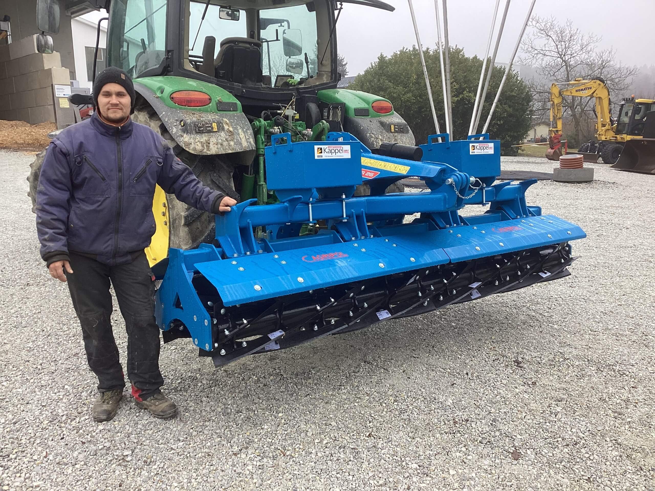 Mann neben Traktor mit angebautem blauen Bodenfräsegerät auf Kiesplatz.